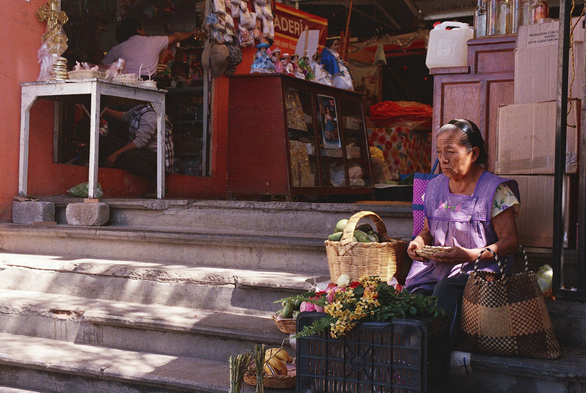 An older woman in an apron sells vegetables and flowers while sitting on the steps of a market in Villa de Etla, Oaxaca.