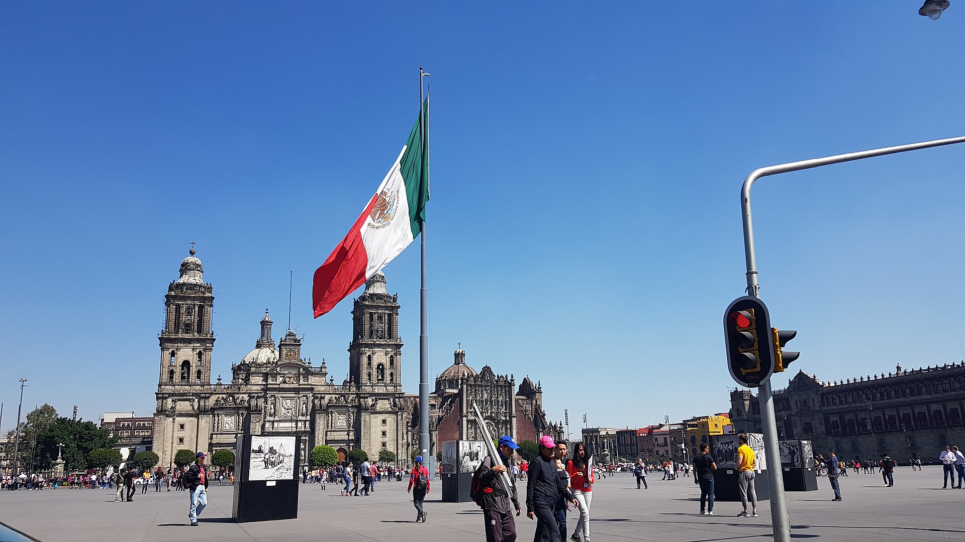 The Mexican flag waves in the Zócalo of Mexico City, in front of the Metropolitan Cathedral and the National Palace, with people walking across the plaza on a sunny day.